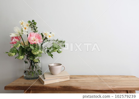 Moody spring still life. Wooden bench, table composition with cup of coffee, tea and old books. Beautiful floral bouquet with white, pink tulips, daffodils. Hawthorn, green guelder rose flowers. Wall. 102579239