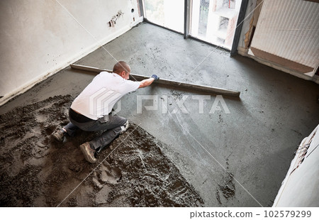 Top view of male construction worker placing screed rail on the floor covered with sand-cement mix. Man smoothing and leveling surface with straight edge while screeding floor in apartment. 102579299