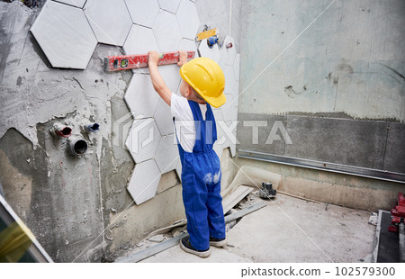 Little boy checking wall surface with spirit level in apartment. Kid in work overalls using level instrument while working on home renovation. 102579300