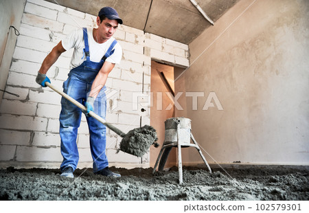 Man preparing floor screed material while standing near concrete screed mixer machine. Male worker using shovel while shoveling sand-cement mix in building under construction. 102579301