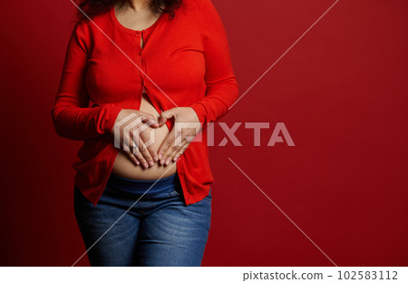 Close-up pregnant woman in casual clothes, makes heart shape with her hands over her bare belly, isolated on red background. Happy carefree pregnancy concept. Enjoying maternity leave. Women's health 102583112