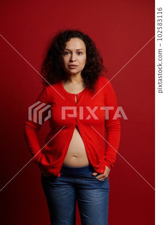 Confident stylish Latin American young pregnant woman wearing unbuttoned red shirt and blue jeans, posing bare belly on red backdrop, looking at camera. Expectant mother. Pregnancy fashion. Maternity Confident stylish Latin American young pregnant woman wearing unbuttoned red shirt and blue jeans, posing bare belly on red backdrop, looking at camera. Expectant mother. Pregnancy fashion. Maternity 102583116