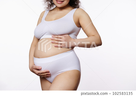 Midsection of a young pregnant woman, expectant gravid mother, putting hands on her bare belly with stretch marks, posing in white underwear over isolated background. Body positivity. Female body 102583135