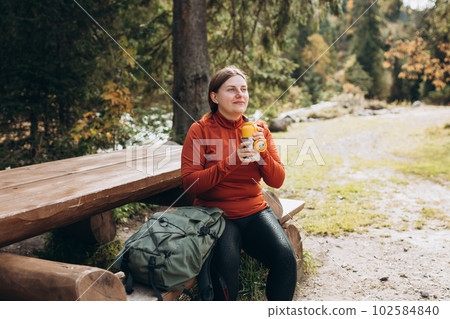 Happy redhead Woman in active trekking clothes having a halt after hiking. Hiker with backpack drinking water from water bottle or hot drink from yellow thermos Happy redhead Woman in active trekking clothes having a halt after hiking. Hiker with backpack drinking water from water bottle or hot drink from yellow thermos 102584840