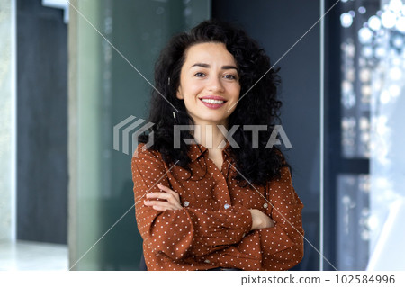Portrait of happy and successful business woman, boss in shirt smiling and looking at camera inside office with crossed arms, Hispanic woman with curly hair in corridor Portrait of happy and successful business woman, boss in shirt smiling and looking at camera inside office with crossed arms, Hispanic woman with curly hair in corridor 102584996