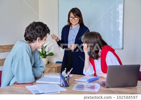 Middle aged female teacher giving lesson to teenage college students sitting at table 102588597