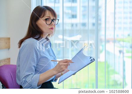Middle aged woman with clipboard sitting in office near window. Middle aged woman with clipboard sitting in office near window. 102588600