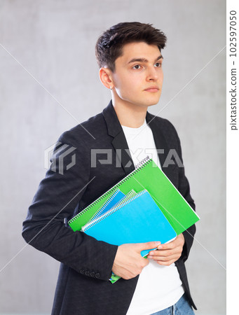 Young male scientist in jacket stands with large stack of notes against gray wall. Young male scientist in jacket stands with large stack of notes against gray wall. 102597050