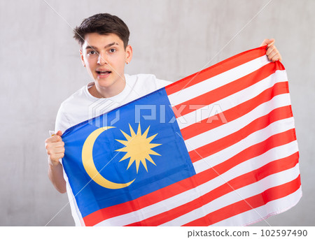 Happy young man holding flag of Malaysia against unicoloured background 102597490