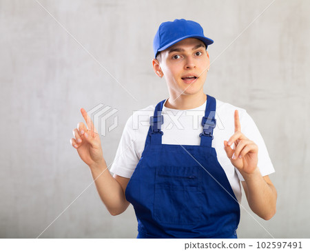 Man in overalls and cap is actively gesticulating-he spread hands to sides,raised index fingers up 102597491