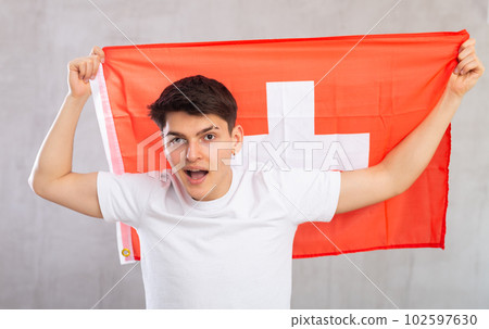 Man in casual clothes hold unfurled flag of Switzerland in hands raised above head against gray wall 102597630