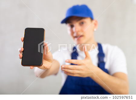 Unfocused man in jumpsuit and cap shows close up empty phone screen and points at device with finger Unfocused man in jumpsuit and cap shows close up empty phone screen and points at device with finger 102597651
