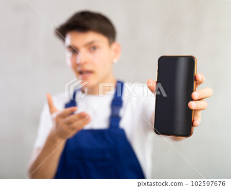 Unfocused man in jumpsuit shows close up empty mobile phone screen and points at device with finger Unfocused man in jumpsuit shows close up empty mobile phone screen and points at device with finger 102597676
