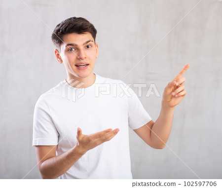 Surprised young man with pointing hand gestures standing against background of light unicolored wall Surprised young man with pointing hand gestures standing against background of light unicolored wall 102597706