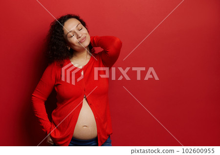 Delightful pregnant woman with her eyes closed, enjoying happy moments of pregnancy and maternity, posing bare belly, isolated on red background. Copy ad space. Expecting baby. Femininity. Sensuality Delightful pregnant woman with her eyes closed, enjoying happy moments of pregnancy and maternity, posing bare belly, isolated on red background. Copy ad space. Expecting baby. Femininity. Sensuality 102600955