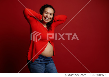 Curly haired ethnic pregnant adult woman laughing, posing bare belly over isolated red background. Enjoying wonderful moments of happy carefree pregnancy. Copy space. Maternity. People. Lifestyle 102600975