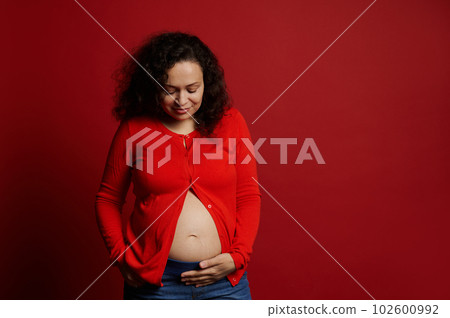 African American positive adult pregnant woman, touching her belly, smiling, enjoying feeling baby movements, isolated on red background with copy advertising space. Pregnancy 28 week. Expecting baby 102600992