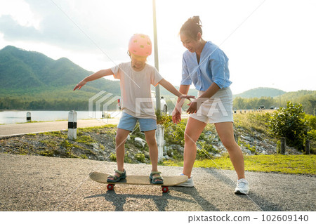 Mother teaching her daughter how to skateboard in the park. Child riding skate board. Healthy sports and outdoor activities for school children in the summer. 102609140