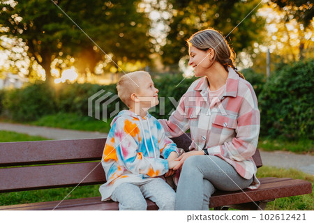 mother and son sit on a park bench in the rays of the setting sun. the concept of a family. Mother's Day. beautiful girl (mother) with a boy (son) in the park in the park are sitting on a bench at mother and son sit on a park bench in the rays of the setting sun. the concept of a family. Mother's Day. beautiful girl (mother) with a boy (son) in the park in the park are sitting on a bench at 102612421