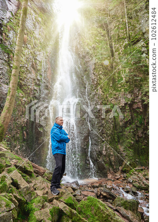 Tourist attraction of Germany - falls of Burgbach Waterfall near Schapbach, Black Forest, Baden-Wurttemberg, Germany. Man hiker in blue jacket standing on stone and looks at flow of falling water 102612484