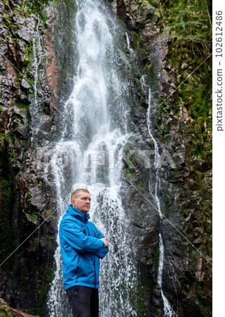 Tourist attraction of Germany - falls of Burgbach Waterfall near Schapbach, Black Forest, Baden-Wurttemberg, Germany. Man hiker in blue jacket standing on stone and looks at flow of falling water 102612486