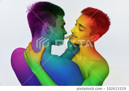 Portrait of two young men, couple hugging, eating jelly candy against white background. Models posing shirtless with rainbow colored body 102613329