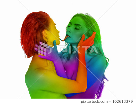 Portrait of loving young women kissing, eating jelly candy against white background. Models painted into rainbow colors. Lgbt 102613379