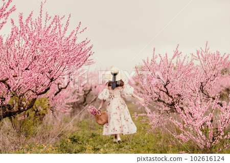 Woman blooming peach orchard. Against the backdrop of a picturesque peach orchard, a woman in a long dress and hat enjoys a peaceful walk in the park, surrounded by the beauty of nature. 102616124