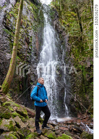 Tourist attraction of Germany - falls of Burgbach Waterfall near Schapbach, Black Forest, Baden-Wurttemberg, Germany. Man hiker in blue jacket standing on stone and looks at flow of falling water Tourist attraction of Germany - falls of Burgbach Waterfall near Schapbach, Black Forest, Baden-Wurttemberg, Germany. Man hiker in blue jacket standing on stone and looks at flow of falling water 102616279