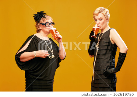 Portrait with two women senior and mature in vintage clothes drinking foamy lager beer over yellow background. Fresh beer draft 102619780