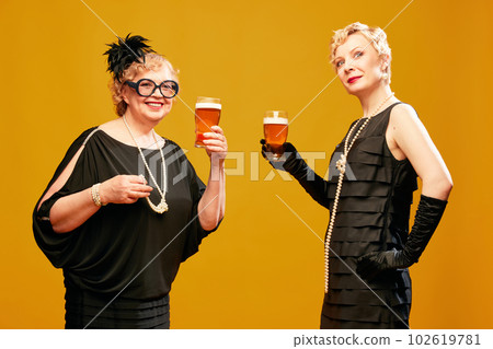 Portrait with attractive two women, friends wearing black dresses enjoying fresh beer over yellow background. Female party 102619781