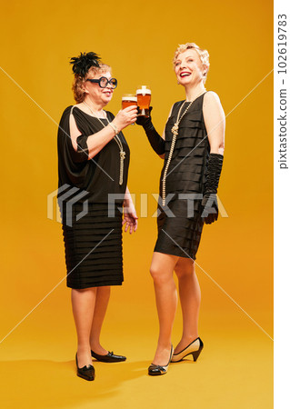 Portrait with two women senior and mature in vintage clothes holding beer glasses and smiling over yellow background. Alcohol degustation 102619783