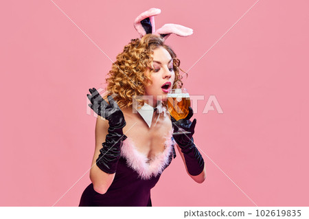 Portrait with adorable young girl, woman wearing bunny costume with ears drinking light beer over pink background. Party, celebrating, ad Portrait with adorable young girl, woman wearing bunny costume with ears drinking light beer over pink background. Party, celebrating, ad 102619835
