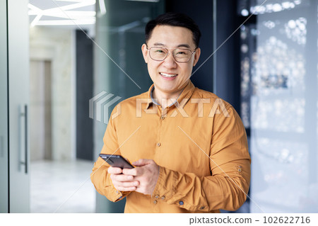 Portrait of a successful Asian man inside the office, a man near window smiling and looking at the camera, a businessman holds a phone in his hands, types a message and browses the Internet. 102622716