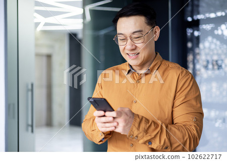 Satisfied with achievement Asian businessman inside office using phone, mature man in shirt near window reading news online from smartphone, successful boss in glasses 102622717