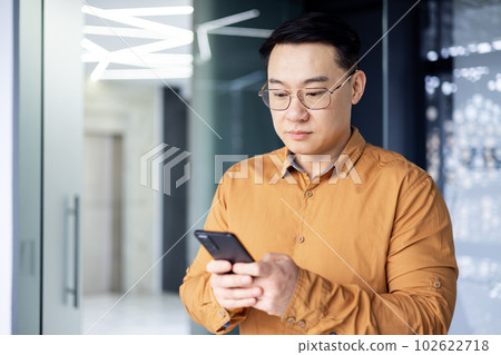 A serious and concentrated Asian man inside the office is using the phone, a businessman is standing by the window browsing the Internet using an application on a smartphone reading news and texting 102622718