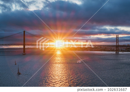 View of 25 de Abril Bridge famous tourist landmark of Lisbon connecting Lisboa and Almada on Setubal Peninsula over Tagus river with tourist yacht boats on sunset. Lisbon, Portugal 102631687