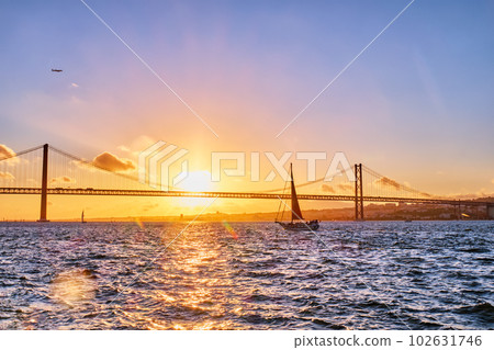 View of 25 de Abril Bridge famous tourist landmark of Lisbon connects Lisboa and Almada on Setubal Peninsula over Tagus river with tourist yacht silhouette at sunset and flying plane. Lisbon, Portugal View of 25 de Abril Bridge famous tourist landmark of Lisbon connects Lisboa and Almada on Setubal Peninsula over Tagus river with tourist yacht silhouette at sunset and flying plane. Lisbon, Portugal 102631746
