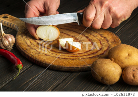 The cook cuts onion with a knife on a cutting board for making soup for lunch. Vegetable diet idea. Work environment on the kitchen table The cook cuts onion with a knife on a cutting board for making soup for lunch. Vegetable diet idea. Work environment on the kitchen table 102631978