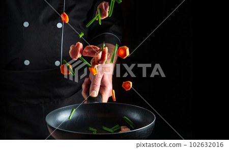 The hand of a professional chef tosses pieces of vegetables and veal meat on a frying pan on a black background. Restaurant cooking concept. Free advertising space The hand of a professional chef tosses pieces of vegetables and veal meat on a frying pan on a black background. Restaurant cooking concept. Free advertising space 102632016