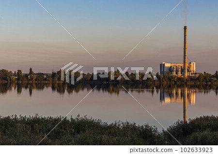 Waste processing plants' chimneys emit smoke at sunset on the lake. Despite generating energy, it leads to air and environmental pollution. Waste processing plants' chimneys emit smoke at sunset on the lake. Despite generating energy, it leads to air and environmental pollution. 102633923