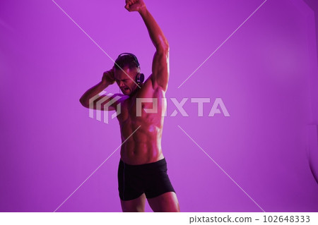 Man wearing headphones athletic physique bodybuilder nude torso abs full-length, fitness classes, purple colored light. Advertising, sports, active lifestyle, competition, challenge concept.  102648333