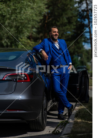 Caucasian bearded man in a blue suit gets out of a black electro car in the countryside in summer. Caucasian bearded man in a blue suit gets out of a black electro car in the countryside in summer. 102657725