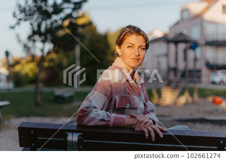 Girl enjoying city view from a bench in sunset or sunrise time. Beautiful blond-haired pretty girl in summer green park against sunset beams. Lonely woman sitting alone moments sunset. On Blurred Girl enjoying city view from a bench in sunset or sunrise time. Beautiful blond-haired pretty girl in summer green park against sunset beams. Lonely woman sitting alone moments sunset. On Blurred 102661274