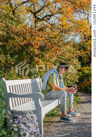 A Teenager Sits On A Bench In The Autumn Park Drinks Coffee From A Thermo Mug And Looks Into A Phone. Portrait Of Handsome Cheerful Guy Sitting On Bench Fresh Air Using Device Browsing Media Smm A Teenager Sits On A Bench In The Autumn Park Drinks Coffee From A Thermo Mug And Looks Into A Phone. Portrait Of Handsome Cheerful Guy Sitting On Bench Fresh Air Using Device Browsing Media Smm 102661344
