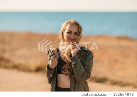 Portrait blonde sea cape. A calm young blonde in an unbuttoned khaki raincoat stands on the seashore, under the raincoat there is a black skirt and top 102664078