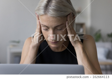 Tired mature blonde woman in casual touching head at laptop Tired mature blonde woman in casual touching head at laptop 102664113