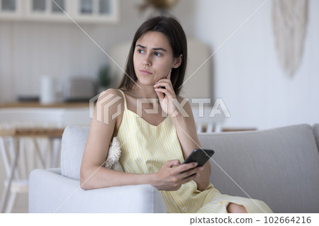 Serious thoughtful young woman holding smartphone oh home couch Serious thoughtful young woman holding smartphone oh home couch 102664216