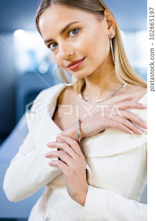 Amazing beautiful model stands in mirror in a jewelry store tries on a gold diamond necklace that sparkles on the girl. Girl is adorned with golden jewelry and diamonds in a luxurious boutique. 102665197