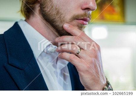 Handsome man in a stylish fashionable suit holds chin at a jewelry store. A male model with a ring on his finger holds face in close-up at a diamond shop. 102665199